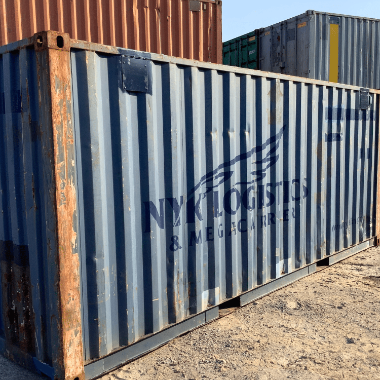 Side view of a blue container labeled “NYK LOGISTICS & MEGACARRIER” in bold letters. The container’s surface is visibly rusted along the edges, and other stacked containers are in the background on a gravel lot.