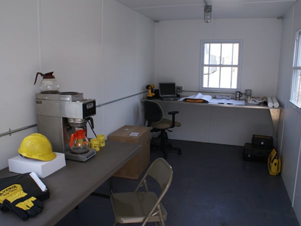 Interior of an A-Verdi office trailer set up at a jobsite, featuring a long desk with a laptop, documents, and tools, as well as a table with a coffee maker, safety gloves, and a hard hat. The space is well-lit with windows on both sides.