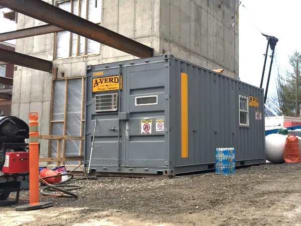 Gray A-Verdi storage container with windows and an air conditioning unit positioned at an active jobsite next to a concrete building structure. The container features yellow A-Verdi logos and a vertical safety stripe, with construction equipment, materials, and caution signs visible nearby.