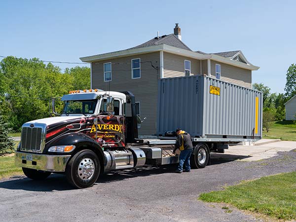 A-Verdi delivery truck placing a gray storage container in the driveway of a two-story home.