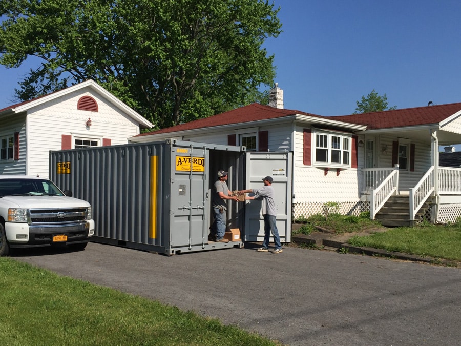 Gray A-Verdi storage container in a residential driveway with two people loading household items for storage.