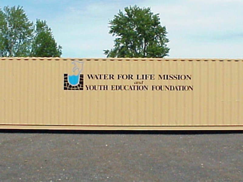 A-Verdi storage container featuring "Water for Life Mission and Youth Education Foundation" branding, set outdoors on a sunny day with trees in the background.