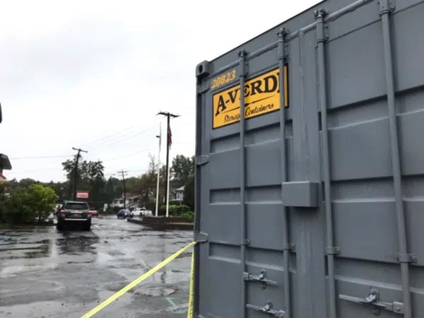 A close-up of a gray A-Verdi storage container with a yellow company logo and identification number 20823, positioned in a wet parking lot on a rainy day. Yellow caution tape runs from the container toward the background, where cars, trees, utility poles, and an American flag are visible.
