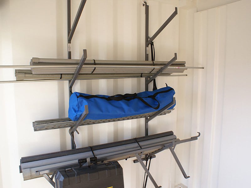 Wall-mounted metal shelving inside a storage container holding pipes, a blue carrying bag, and folded equipment.