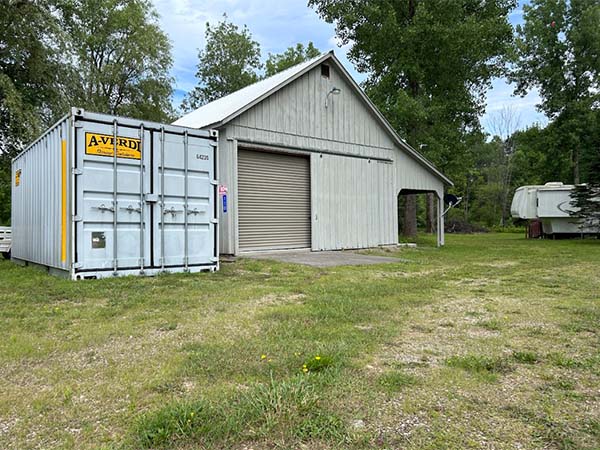Gray A-Verdi storage container positioned beside a large light gray barn with a roll-up door.