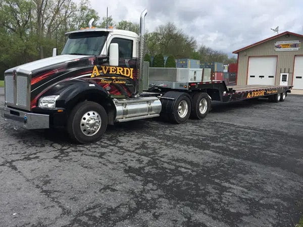 Black A-Verdi semi-truck with bold red and yellow graphics parked in front of a building, attached to a flatbed trailer loaded with equipment. The truck is positioned on a paved lot under a cloudy sky.