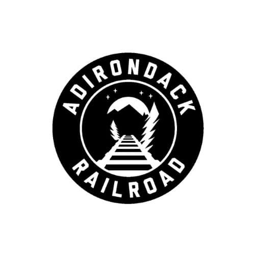 A bold, black, circular logo with the words "ADIRONDACK RAILROAD" in uppercase letters wrapping around the outer edge. Inside, a stylized railroad track leads toward a mountain peak, framed by tall pine trees and small stars in the sky.