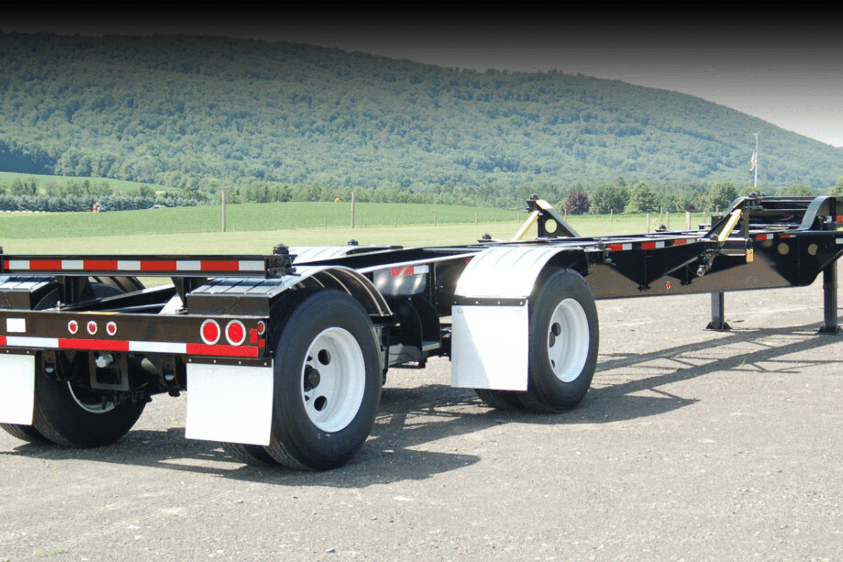 Close-up view of a black and white container chassis trailer parked on a paved lot, designed for transporting shipping containers, with green hills and farmland in the background.