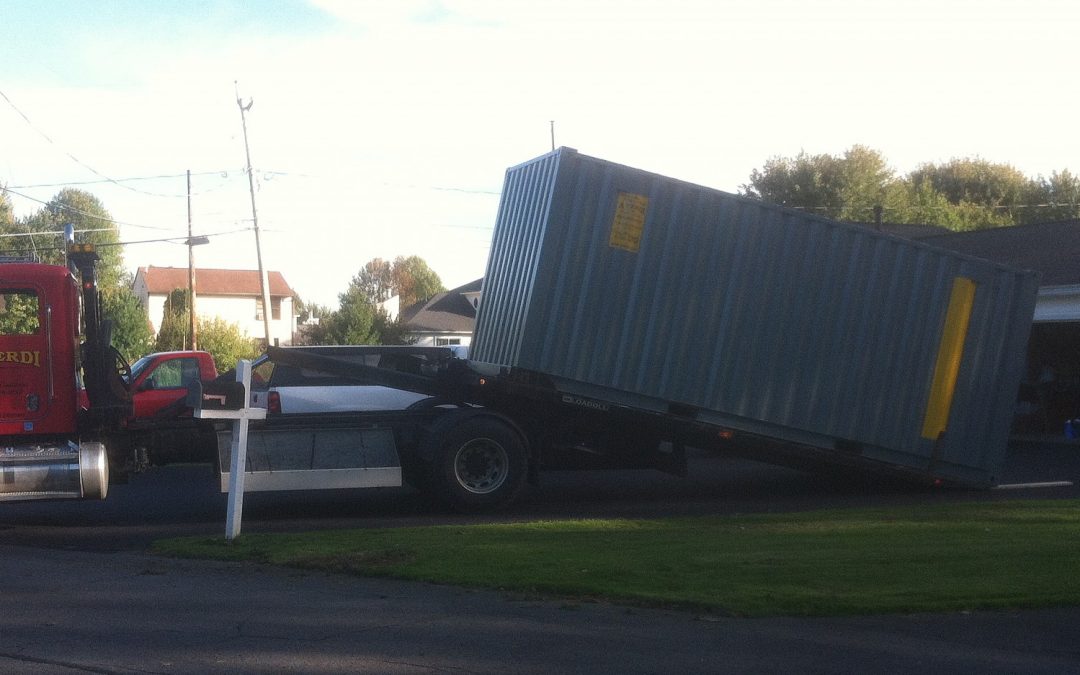 A-Verdi red delivery truck unloading a gray storage container at a residential driveway, positioned near a garage.