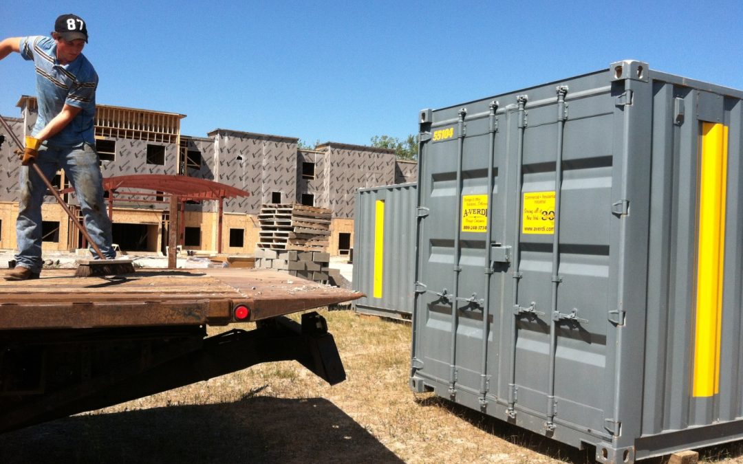 Worker unloading a flatbed truck near storage containers at a construction site with partially built structures in the background.