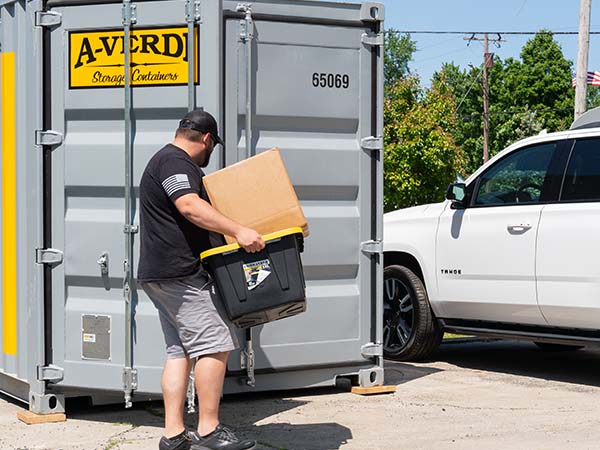 Man carrying a cardboard box and plastic storage bin toward a gray A-Verdi storage container parked beside a white SUV.