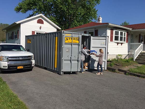 Gray A-Verdi storage container in a residential driveway with two people unloading boxes.