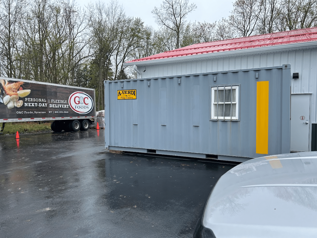 A gray A-Verdi storage container with yellow accents and windows is placed on a paved lot next to a building with a red metal roof. A G&C Foods refrigerated delivery truck is parked nearby, with orange safety cones placed along the driveway. The background features tall trees under an overcast sky.