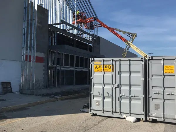 Gray A-Verdi storage containers positioned at an active jobsite in front of a commercial building under construction, with workers installing steel framing from an orange boom lift. The yellow A-Verdi logos are clearly visible under a sunny blue sky.