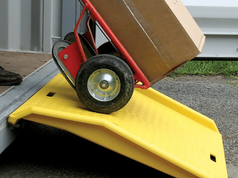 Hand truck with a large box being rolled up a bright yellow portable loading ramp into a storage container.