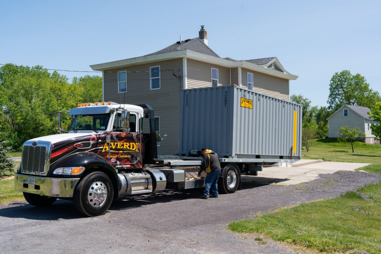 A-Verdi truck delivering a gray storage container to a residential driveway, with a worker securing it.